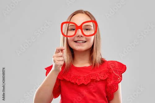 party props  photo booth and childhood concept - beautiful smiling girl in red shirt with big paper glasses over grey background