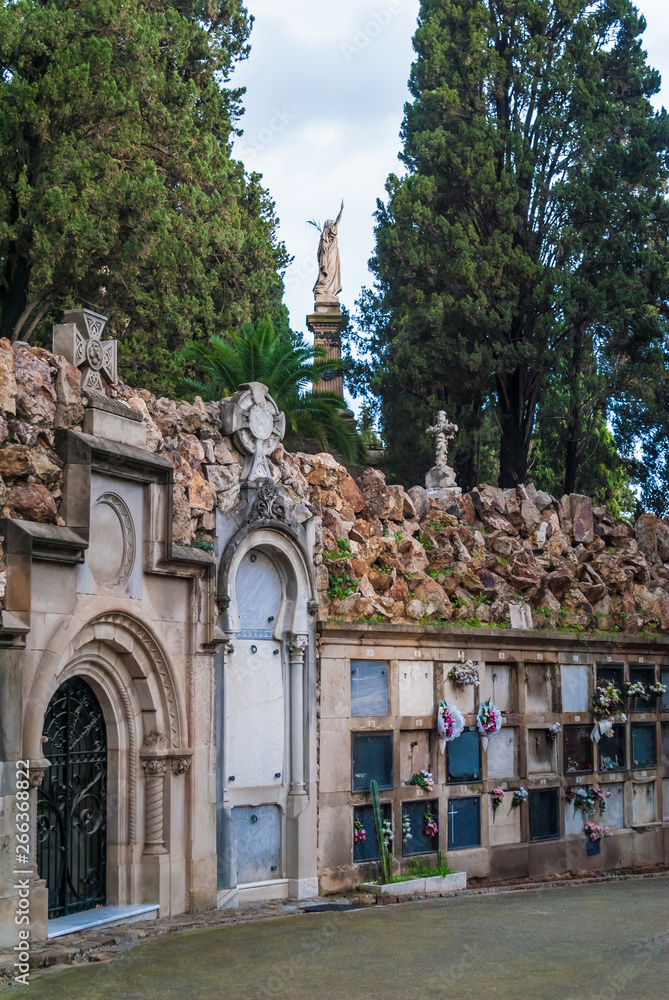 The stone wall with graves and crypt and high sculpture behind it on the Montjuic Cemetery in overcast day, Barcelona, Catalonia, Spain
