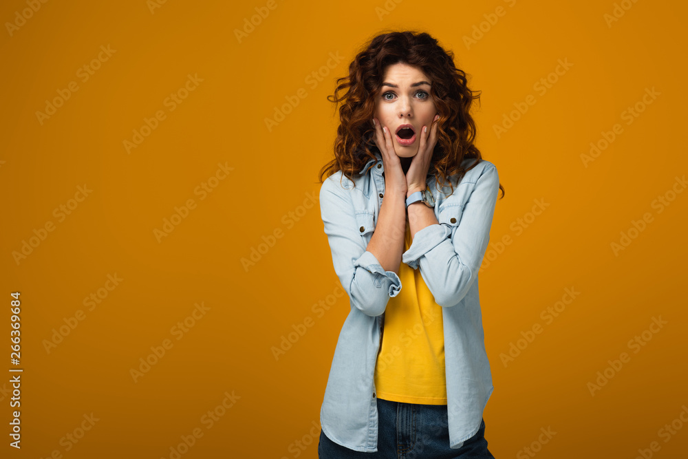 surprised redhead woman looking at camera and touching face on orange surprised redhead woman looking at camera and touching face on orange