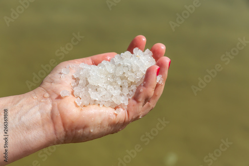 Middle age woman holding salt crystals in her hand with the Dead Sea in the Background during a bright sunny day. Taken in Ein Bokek, Israel.