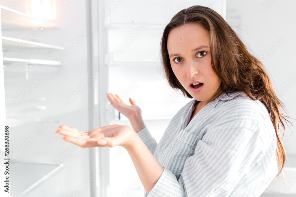 Surprised girl standing in the kitchen looks into the empty refrigerator spreading his hands to the sides. Out of food Surprised girl standing in the kitchen looks into the empty refrigerator spreading his hands to the sides. Out of food