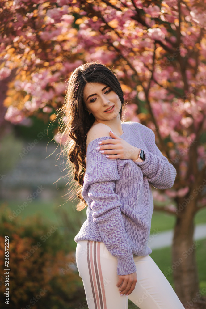 Attractive young woman in a purple sweater stand in front of sakura tree in the park Attractive young woman in a purple sweater stand in front of sakura tree in the park