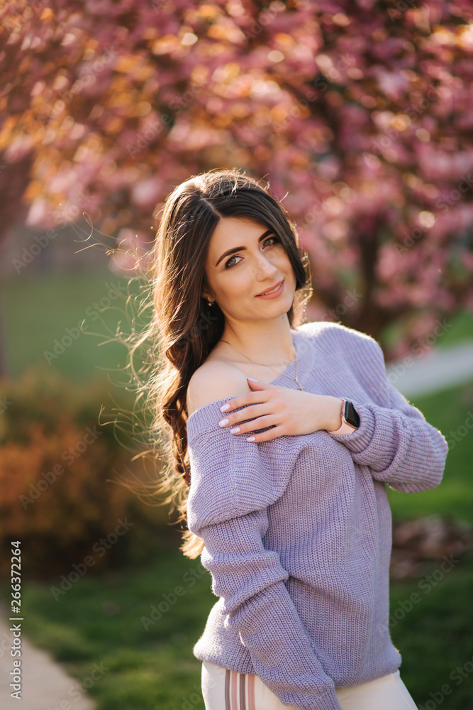 Attractive young woman in a purple sweater stand in front of sakura tree in the park Attractive young woman in a purple sweater stand in front of sakura tree in the park