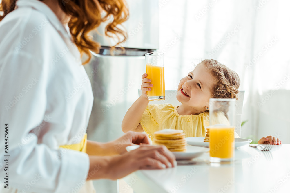 selective focus of emotional happy daughter looking at mother and holding glass with orange juice while having breakfast selective focus of emotional happy daughter looking at mother and holding glass with orange juice while having breakfast