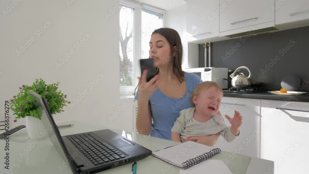 combining work and parenting, mother with crying little son while working on laptop computer and talking on mobile phone sitting at table in kitchen at home combining work and parenting, mother with crying little son while working on laptop computer and talking on mobile phone sitting at table in kitchen at home