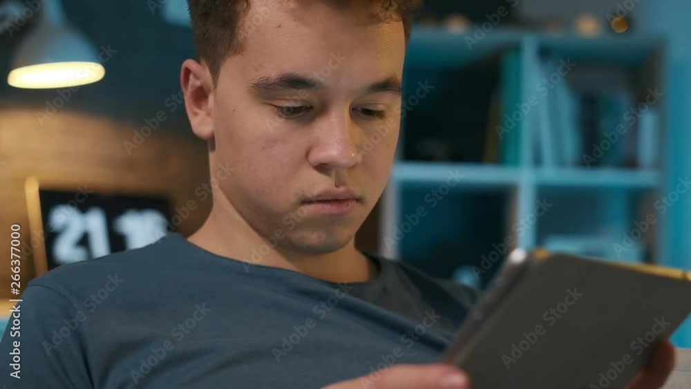 Close-up view of a boy attentively looking at his tablet while sitting on a couch in his room Close-up view of a boy attentively looking at his tablet while sitting on a couch in his room