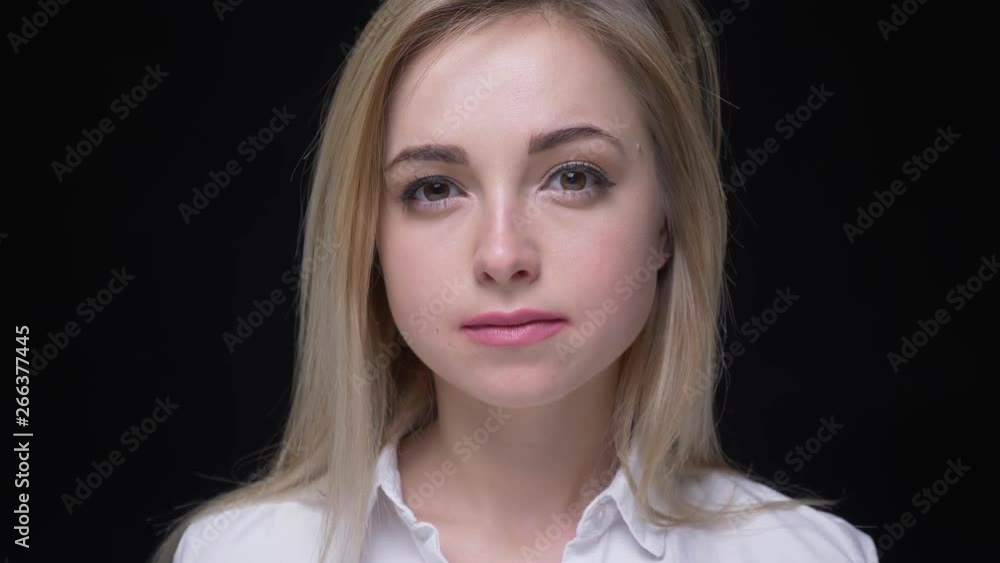 Close-up shoot of pretty young blonde girl with big brown eyes in white shirt watches attentively on black background.