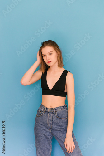 Beautiful model in a bra and jeans poses on a blue background, looking into the camera. Portrait of a woman with natural light on a blue background. Tests for a model agency. Model's snapshots.