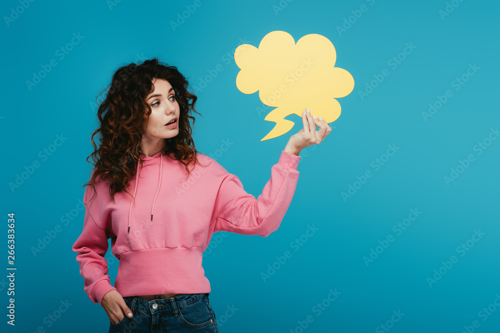 attractive curly redhead girl holding thought bubble while standing with hand in pocket on blue attractive curly redhead girl holding thought bubble while standing with hand in pocket on blue