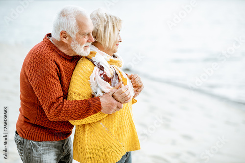 Lovely senior couple dressed in colorful sweaters hugging on the sandy beach, enjoying free time during retirement near the sea #266386668