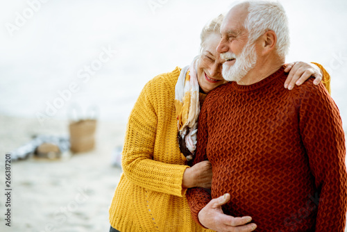 Portrait of a happy senior couple dressed in colorful sweaters hugging on the sandy beach, enjoying free time during retirement near the sea photo