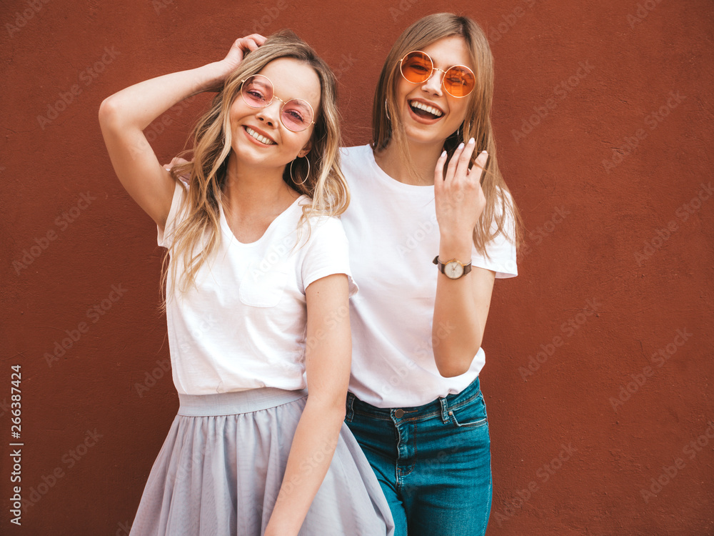 Two young beautiful blond smiling hipster girls in trendy summer white t-shirt clothes. Women posing in the street near red wall. Positive models having fun in sunglasses