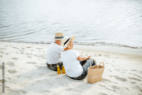 Senior couple sitting together with drinks and bag on the sandy beach, enjoying their retirement near the sea, rear view
