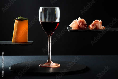 Composition of red wine in glass and pieces of prosciutto and cheese snack on black background