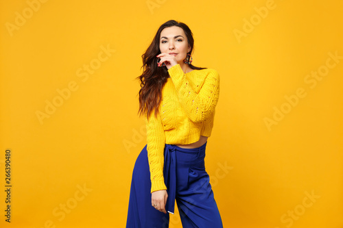 Portrait of attractive young woman in sweater, blue trousers looking camera rising hand isolated on yellow orange background in studio. People sincere emotions, lifestyle concept. Mock up copy space.