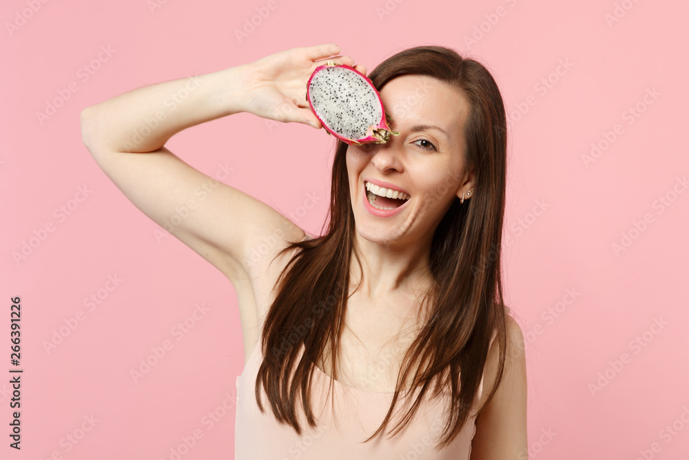 Cheerful young woman covering eye with half of fresh ripe pitahaya, dragon fruit isolated on pink pastel wall background in studio. People vivid lifestyle relax vacation concept. Mock up copy space. Cheerful young woman covering eye with half of fresh ripe pitahaya, dragon fruit isolated on pink pastel wall background in studio. People vivid lifestyle relax vacation concept. Mock up copy space.