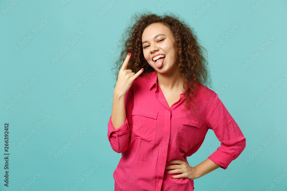 Obraz premium Cheerful african girl in pink casual clothes showing tongue, depicting heavy metal rock sign isolated on blue turquoise wall background. People sincere emotions lifestyle concept. Mock up copy space.