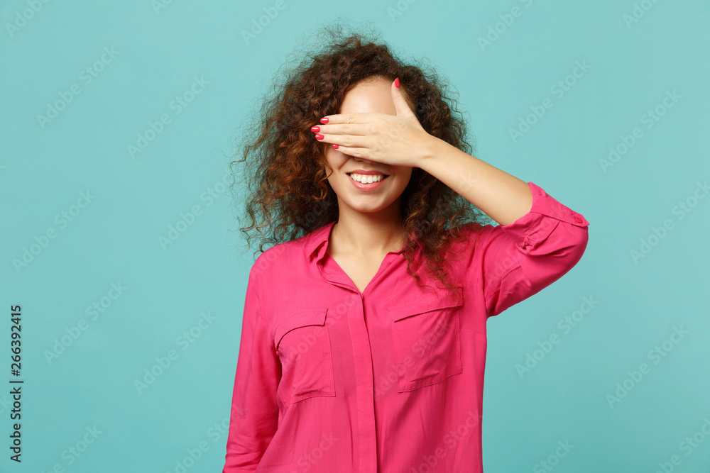 Portrait of smiling african girl in pink casual clothes covering eyes with hand isolated on blue turquoise wall background in studio. People sincere emotions, lifestyle concept. Mock up copy space. Portrait of smiling african girl in pink casual clothes covering eyes with hand isolated on blue turquoise wall background in studio. People sincere emotions, lifestyle concept. Mock up copy space.