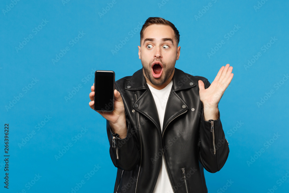 Young man in black jacket white t-shirt hold mobile phone with blank empty screen for promotional content isolated on blue wall background studio portrait. People lifestyle concept. Mock up copy space Young man in black jacket white t-shirt hold mobile phone with blank empty screen for promotional content isolated on blue wall background studio portrait. People lifestyle concept. Mock up copy space