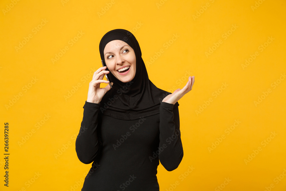 Smiling young arabian muslim woman in hijab black clothes hold in hands talking on mobile phone isolated on yellow background, studio portrait. People religious lifestyle concept. Mock up copy space. Smiling young arabian muslim woman in hijab black clothes hold in hands talking on mobile phone isolated on yellow background, studio portrait. People religious lifestyle concept. Mock up copy space.