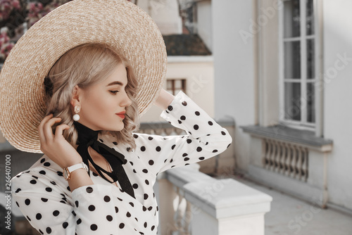 Outdoor fashion portrait of young beautiful lady wearing  trendy pearl earrings, stylish straw wide brim hat with ribbon tie, polka dot blouse, white wrist watch, posing in street. Copy, empty space