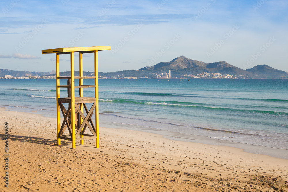 a yellow rescue tower on an empty sand beach of Mediterranean sea bay; city and mountain on the background; Mallorca island in winter.