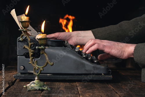 A writer is typing a text on a typewriter on his desk on a burning fire in fireplace background.