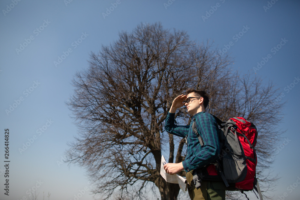 A traveler with a backpack, looking at the map and walking in the countryside. Tree in the background