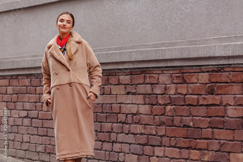 young stylish cute girl in a fur coat strolling around the city near wooden houses and stone walls