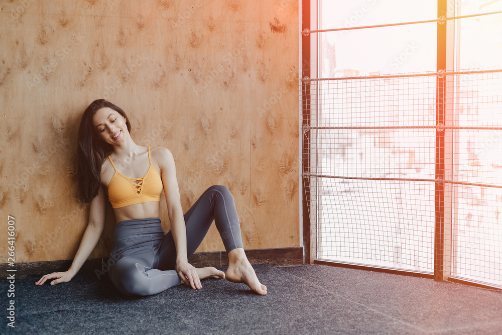 Fototapeta premium Young attractive fitness girl sitting on the floor near the window on the background of a wooden wall, resting on yoga classes