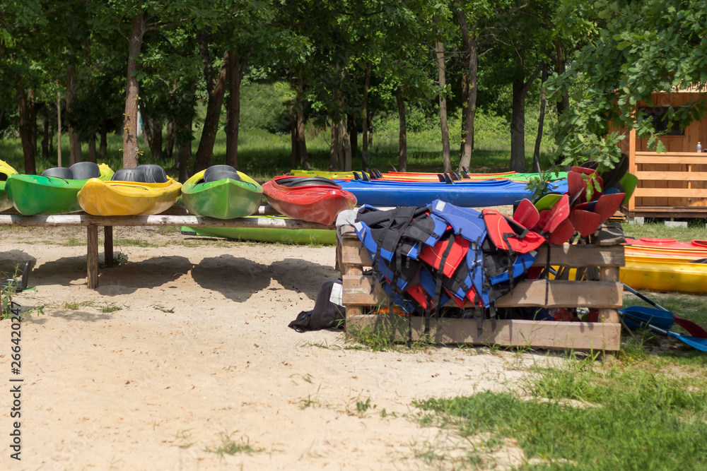 Wypożyczalnia kajaków. Kolorowe kajaki poustawiane na stojakach leżą na plaży. Kapoki i wiosła poukładane w drewnianym pojemniku.