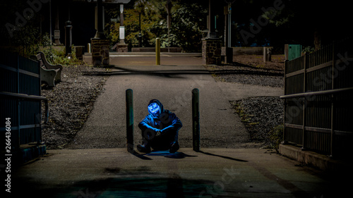 Hooded figure with neon mask blocking path. 