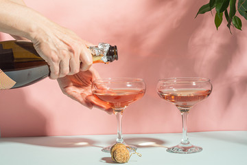Female hand pouring champagne or wine into glasses. Gentle pink background bright sunlight. Copy space minimalism.