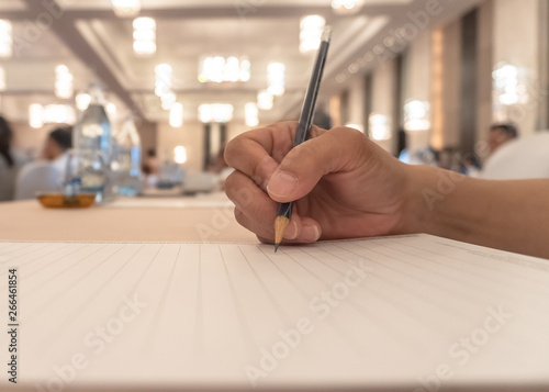 Business agenda briefing shorthand note in seminar workshop or convention hall meeting room with hand holding pencil on notepad for taking note on desk and blur people background photo