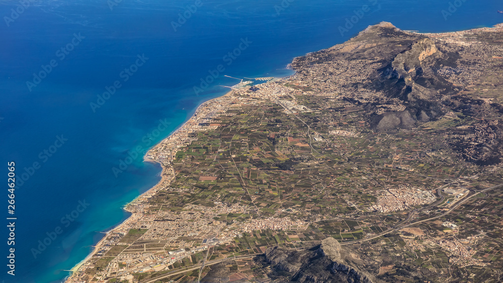 A high-altitude shot of an airplane shows the eastern Spanish city of Denia and the surrounding area with mountains and coastline in sunny weather and blue skies. A high-altitude shot of an airplane shows the eastern Spanish city of Denia and the surrounding area with mountains and coastline in sunny weather and blue skies.