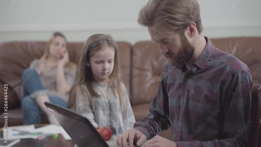 Portrait of the bearded man working with tablet close up. Little girl unsuccessfully trying to give the man the apple while mother sitting on the sofa on the background. Young man addicted to gadgets Portrait of the bearded man working with tablet close up. Little girl unsuccessfully trying to give the man the apple while mother sitting on the sofa on the background. Young man addicted to gadgets