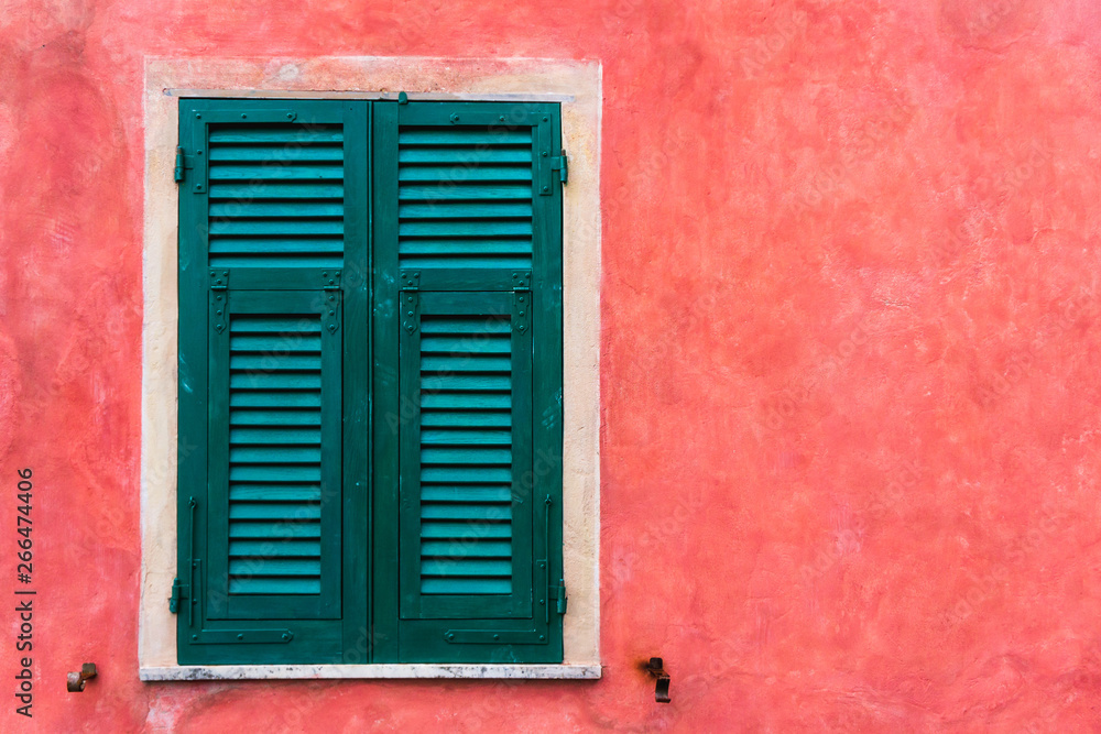 Closed vintage Italian window with exterior green shutters on a textured red wall. 