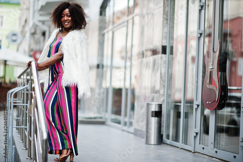 Fashionable african american woman in pink striped jumpsuit with fluffy faux fur coat posed at street of megapolis.