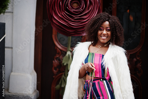 Fashionable african american woman in pink striped jumpsuit with fluffy faux fur coat and handbag posed at street against large rose.