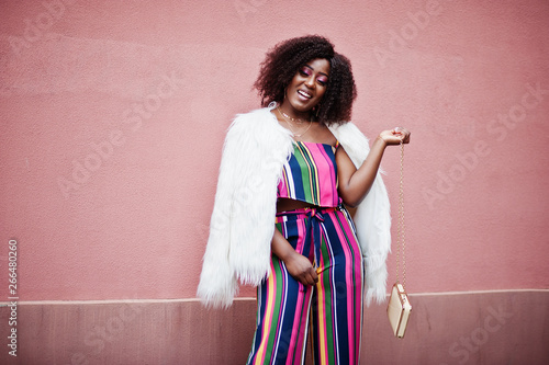 Foto Fashionable african american woman in striped jumpsuit with fluffy faux fur coat and handbag posed against pink wall