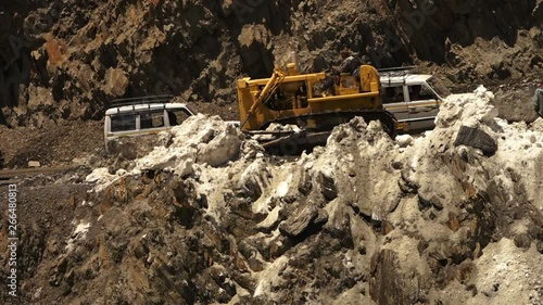 Close up, pan shot of a yellow bulldozer, removing accumulated glaciers from mountain road, ZojiLa Pass, and pushing them down a steep slope. Off-road vehicles are passing heading uphill photo