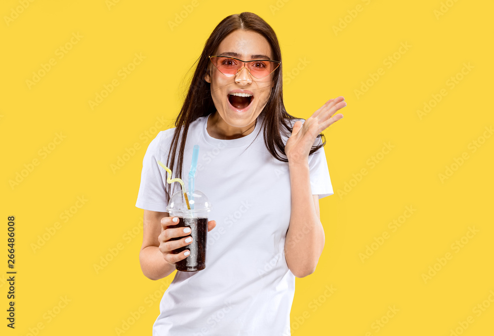 Beautiful female half-length portrait isolated on yellow studio background. Young smiling woman in red sunglasses with drink. Facial expression, summer, weekend, resort concept. Trendy colors. Beautiful female half-length portrait isolated on yellow studio background. Young smiling woman in red sunglasses with drink. Facial expression, summer, weekend, resort concept. Trendy colors.