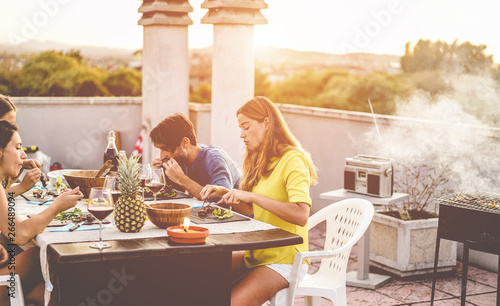 Young friends having barbecue meal at sunset on rooftop patio - Happy people doing bbq dinner outdoor cooking meat and drinking wine - Focus on right woman face - Food, fun and friendship concept