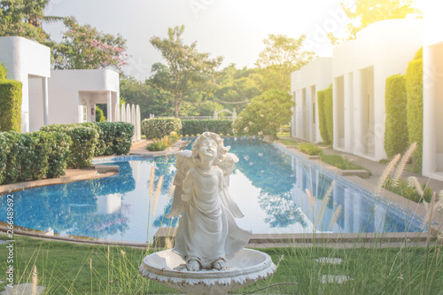 Old white cupid sculpture or statue standing in outdoor garden surrounded with green natural for decoration with sunlight and swimming pool in the background.