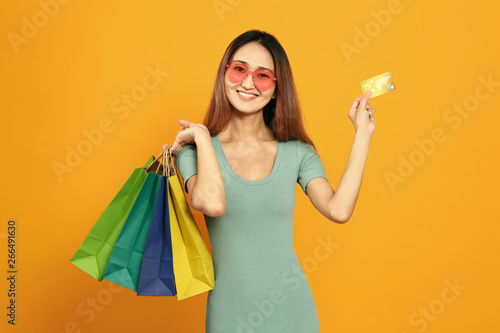 Asian woman with shopping bags and credit card on color background