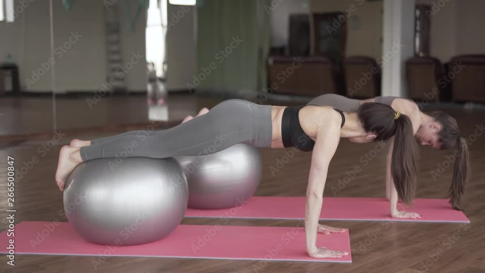 Two young beautiful women in a fitness room with a glass wall are doing an excercise with a fit ball together. Women are very fit and slender. Two young beautiful women in a fitness room with a glass wall are doing an excercise with a fit ball together. Women are very fit and slender.