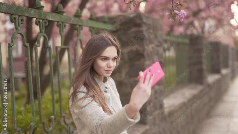 Young Caucasian Female in Gary Jacket Standing By Fence of Park on Street and Taking Selfies With Smartphone. Light Camera Movement Young Caucasian Female in Gary Jacket Standing By Fence of Park on Street and Taking Selfies With Smartphone. Light Camera Movement