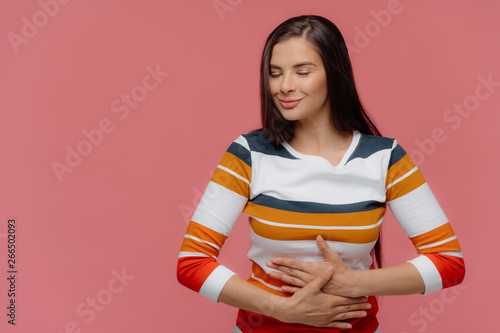 Satisfied woman keeps hands on belly, feels replete after delicious supper, has full stomach, long hair, appealing appearance, wears jumper, models on pink wall. Lady finds out about pregnancy