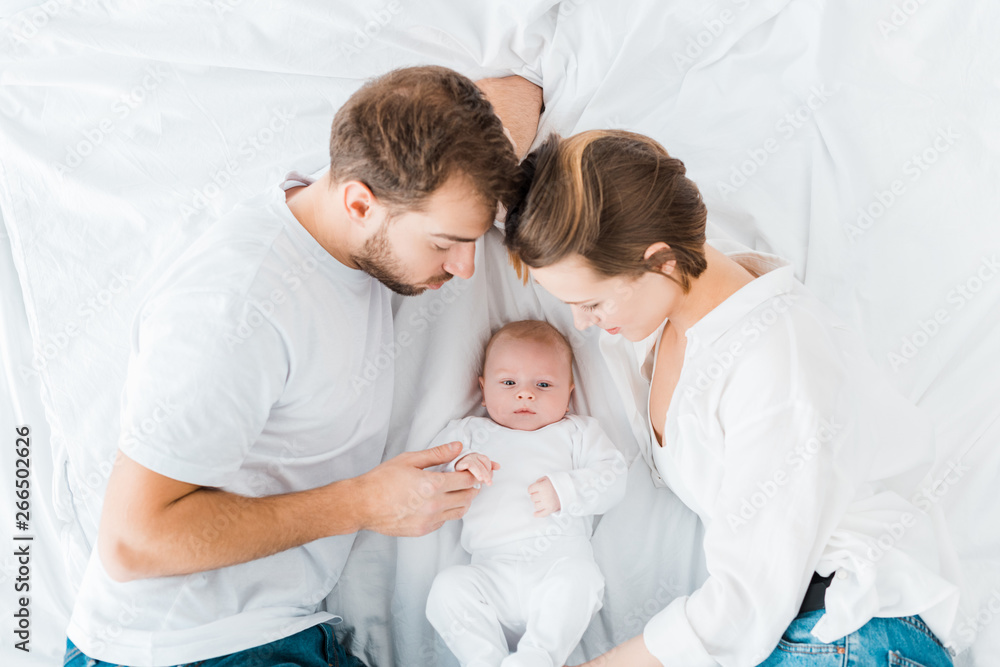 overhead view of happy parents lying on white sheet and looking at baby overhead view of happy parents lying on white sheet and looking at baby