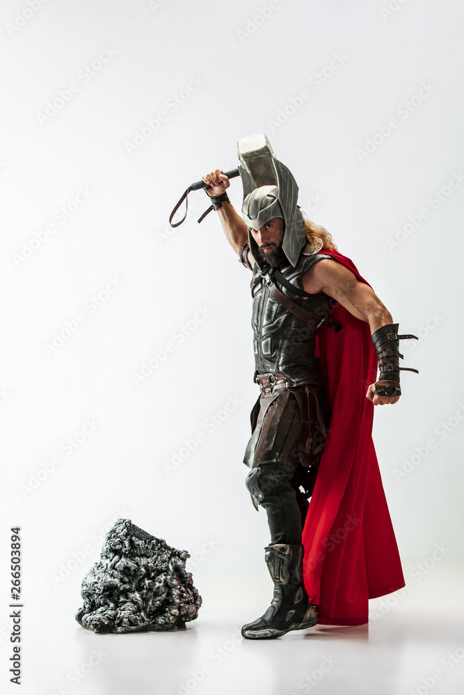 Fototapeta premium Long hair and muscular male model in leather viking's costume with the big hammer cosplaying Thor isolated on white studio background. Full-lenght portrait. Fantasy warrior, antique battle concept.